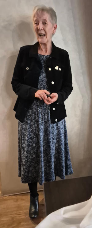 A mgrey-haired lady wearing a black jacket over a blue and white patterned dress