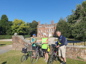 Three people standing by the low wall of a stately home, holding bikes