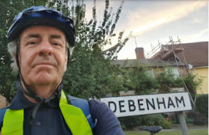 Head and shoulders of a man wearing a cycling helmet standing near a sign reading Debenham